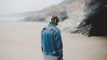 a woman in a denim jacket walking on the beach 