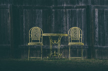 Two yellow wrought-iron chairs and cafe table along wooden fence.