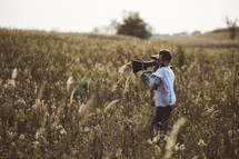 a man with a video camera standing in a field of dried flowers 