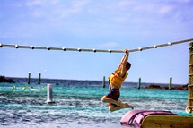 Boy hanging from over-head rope ladder off pier in ocean.