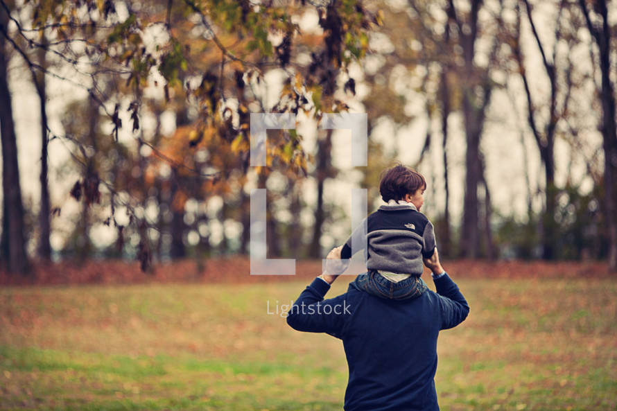 Father walking in tree-lined park with son riding on his shoulders.