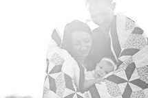Couple holding infant daughter while all wrapped in a quilt.