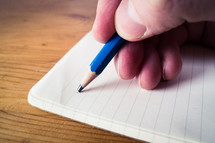 A male hand writing with a blue pencil on a blank open notebook