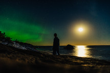 a man walking along a shore at night looking up at the Aurora Borealis 