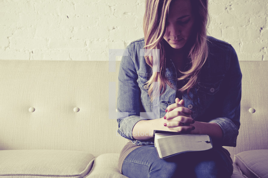 woman sitting on a couch with her hands in prayer held over a Bible.