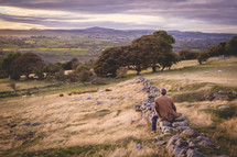 a man sitting on a rustic rock wall on a hillside 