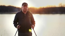 Senior man stand with trekking poles in their hands on the lake, resting after Nordic walking, enjoy nature at sunset time.
