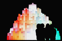 silhouettes standing in front of a glowing rainbow sculpture 