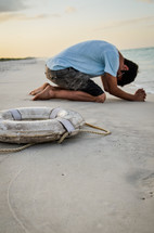 man kneeling in prayer on a beach and a life ring