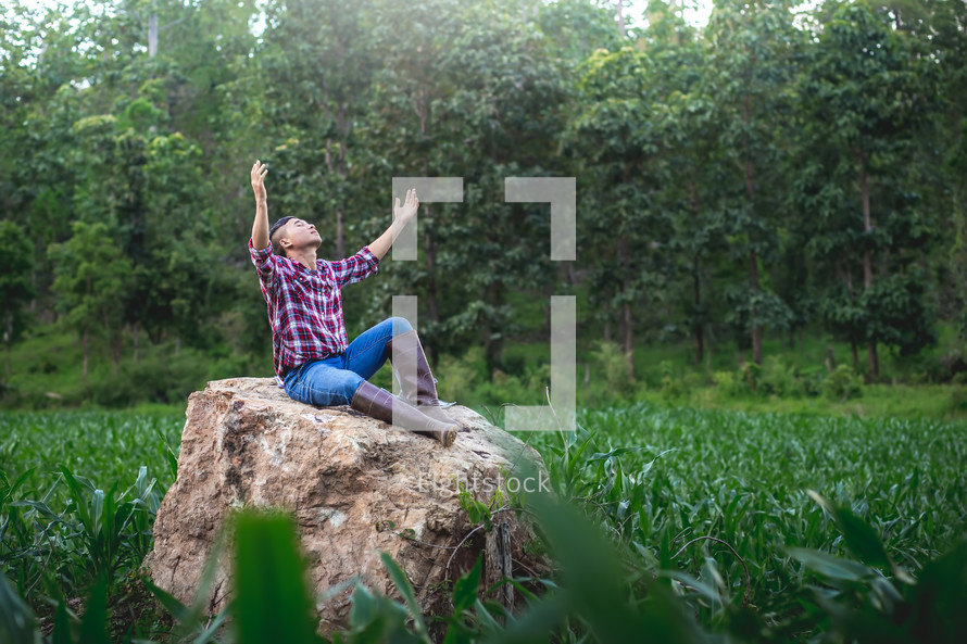 Young farmer sitting on a big stone with hands raised praising God 
