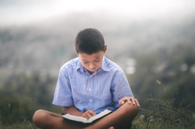 a boy sitting outdoors reading a Bible 