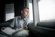 a boy reading a Bible and looking out a window 