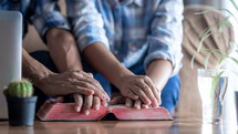 closeup of two people with hands on a Bible praying at home 
