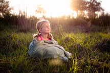 a little girl wrapped in a blanket sitting in tall grasses at sunset 