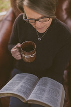 A woman sitting in a chair reading her Bible and drinking coffee