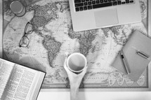 open Bible, mug, reading glasses, laptop, pen, journal, and ink on a world map 