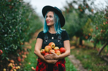 Blue haired girl picked up a lot of ripe red apple fruits from tree in green garden. Organic lifestyle, agriculture, gardener occupation.