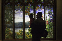 a father holding his toddler looking at a stained glass window 