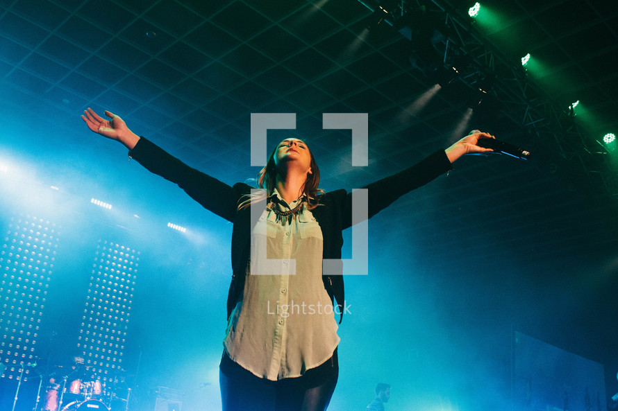 A woman standing on stage with arms outstretched in worship.