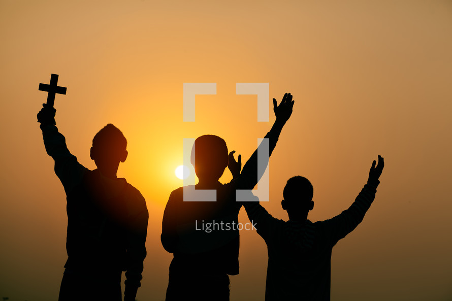 silhouettes of three children standing outdoors at sunset holding a cross