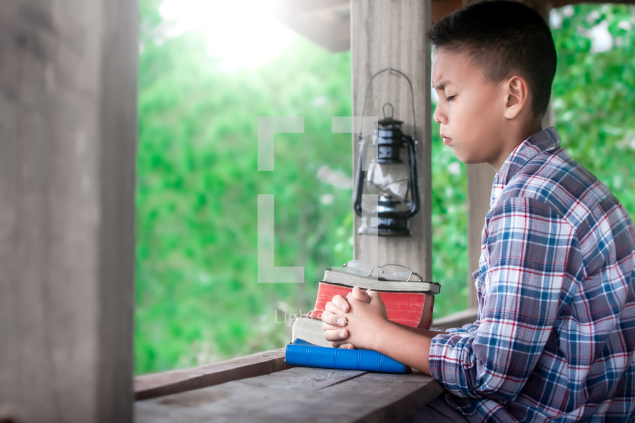 a boy praying outdoors over a stack of Bibles 