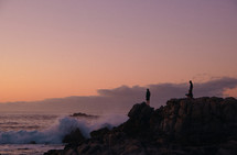 people standing on a rocky shore at sunset watching the waves 