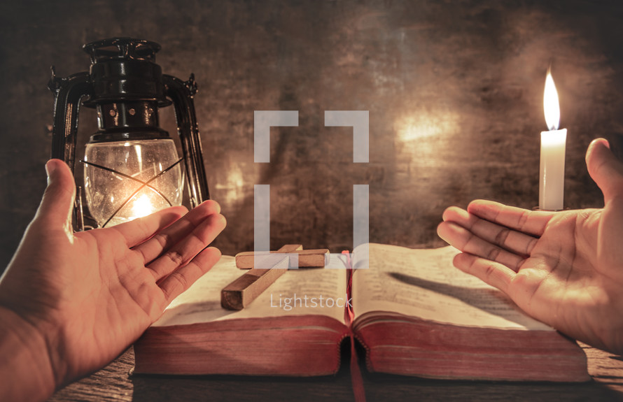 praying hands and cross on an open Bible and light from an oil lamp 