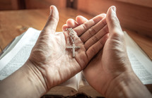 cross necklace in open hands in prayer over a Bible 