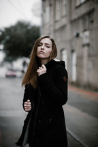 a young woman in a coat standing on a city street looking back at the camera 