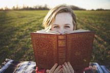 Girl sitting on blanket in grassy field reading ornate William Shakespeare book.