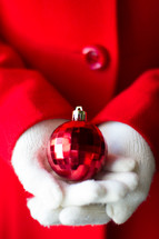 A woman's hands wearing white gloves holding a red Christmas ball 