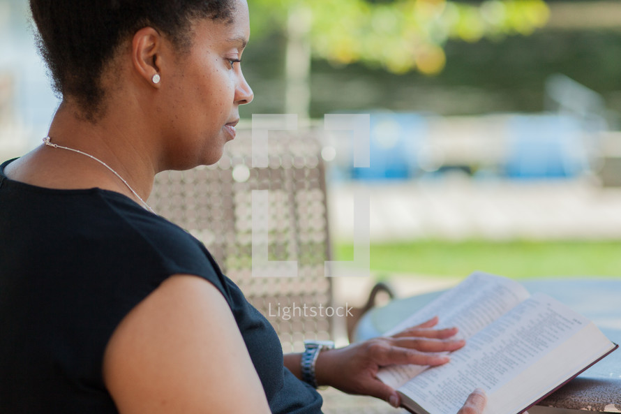 a woman sitting at an outdoor table and chairs reading a Bible 
