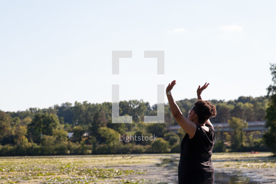 an African American woman with raised hands standing on a lake shore 