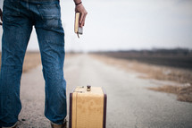 man holding a Bible standing next to a suitcase looking down a long road