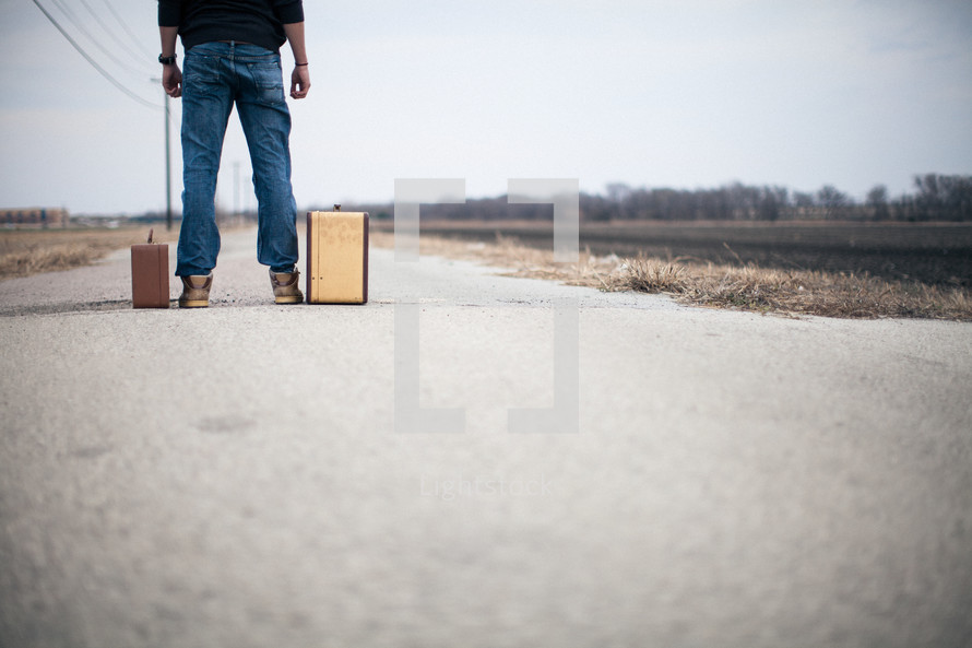 man looking down a road standing next to suitcases