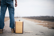 man looking down a road standing next to a suitcase