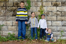 Four children holding hands and standing by a stone wall.