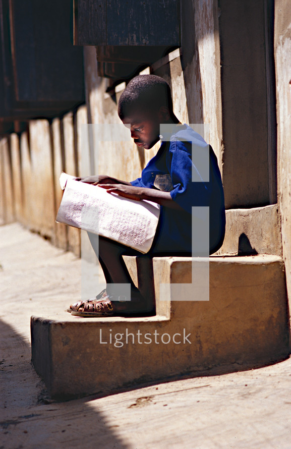 Blind orphan boy sitting on concrete stairs reading a book in Braille 
