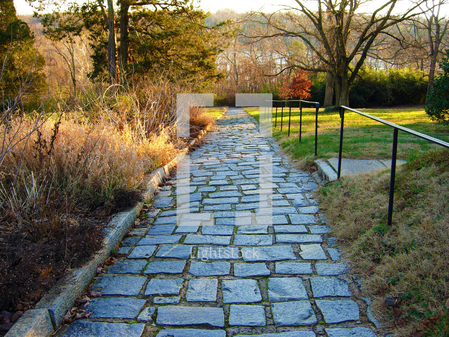 A pathway made out of stones with a metal railing lined with sun drenched grass and trees in a Virginia park outdoors. 