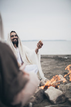 Jesus breaking bread with his disciples around a campfire on the beach.