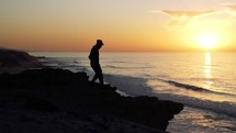 Man in hat walk on rocky beach and looking on beautiful sunset sky above ocean landscape nature
