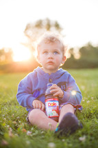 A little boy sitting in green grass with a bottle of juice.