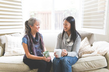 Asian women sitting on a couch talking and holding mugs 