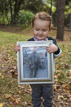 Boy standing in the woods holding a framed photo of his soldIer father.