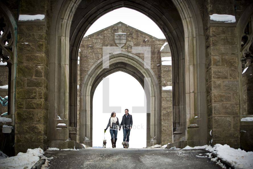 A couple walking dogs through the archways of a building.