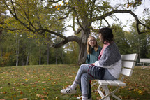 Two young women sitting and talking, smiling on a bench with lovely background