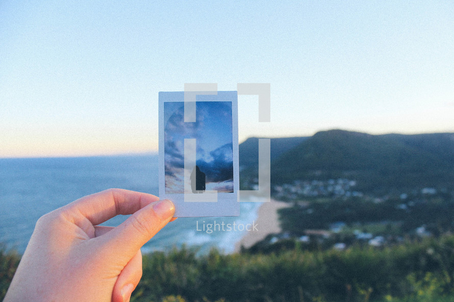 woman holding up a polaroid picture near a coastline 