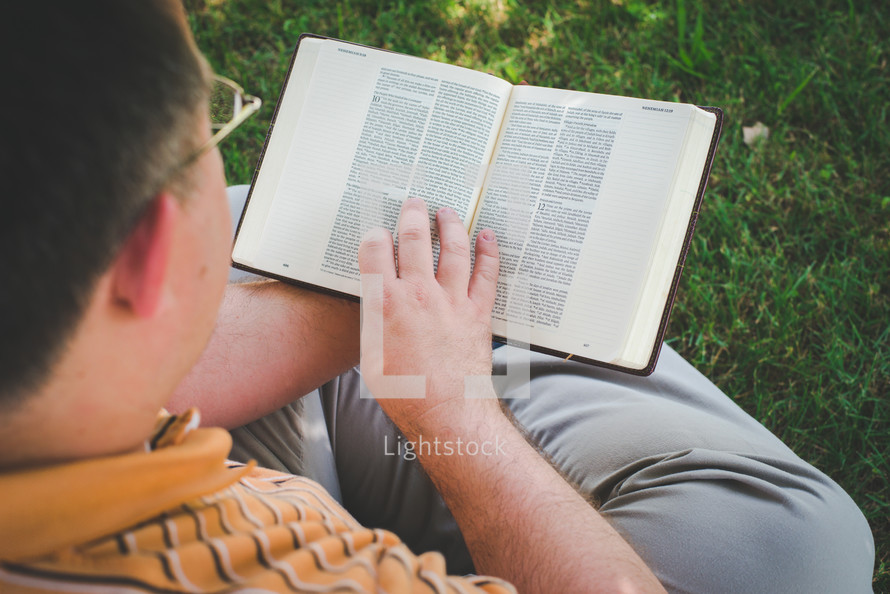 a man sitting in the grass on a sunny day reading a Bible God's Word with a view of him
from above. 
