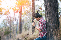 a man sitting under a tree holding a cross praying in a forest 