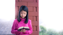 a girl in a pink jacket standing outdoors reading a Bible 